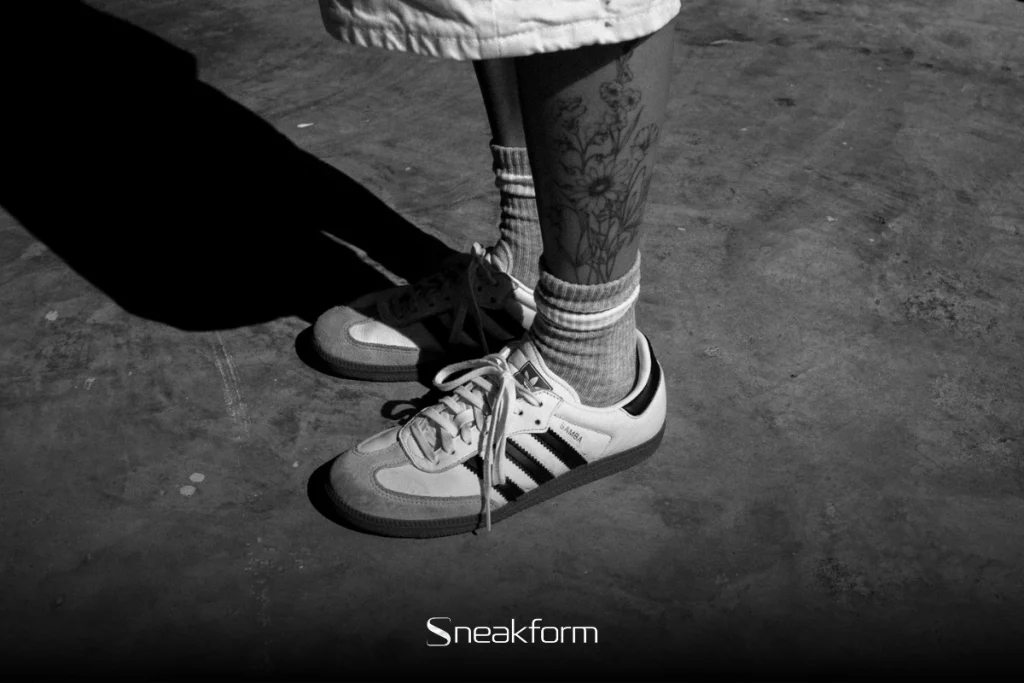 Gros plan en noir et blanc sur une paire d'Adidas Samba OG portée avec des chaussettes côtelées et un tatouage floral sur la cheville, symbole du glissement des sneakers de la rue vers la mode.